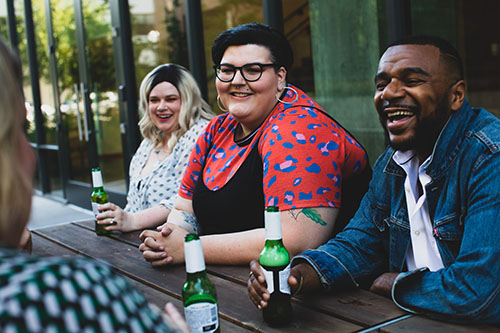 A group of young people laughing and having a conversation over beers at an outdoor picnic table.