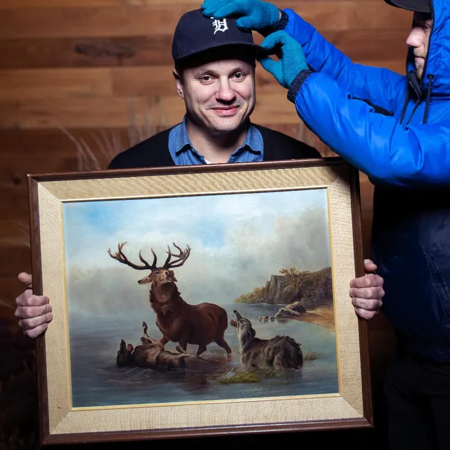 Jim Bryson holding a picture of a stag while a man in a blue coat adjusts Jim's hat.