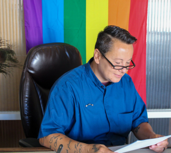 Reverend Jenn sitting at a desk in front of a rainbow pride flag.